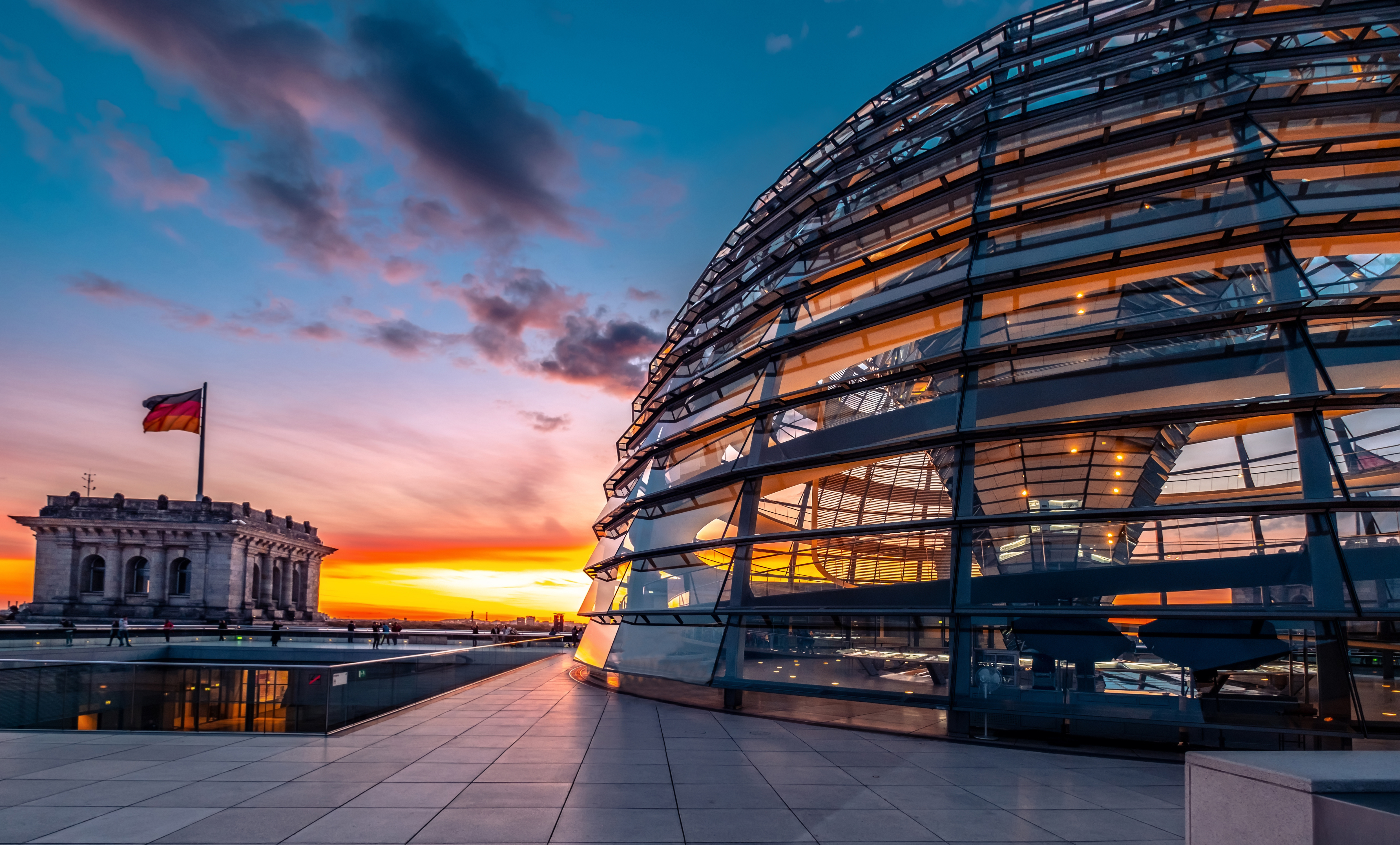 beleuchtete Glaskuppel des Reichstagsgebäudes in Berlin bei Sonnenuntergang, mit einer deutschen Flagge im Hintergrund