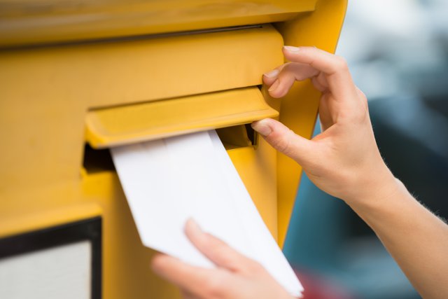 Foto: Ein weißer Briefumschlag wird in einen gelben Briefkasten eingeworfen.