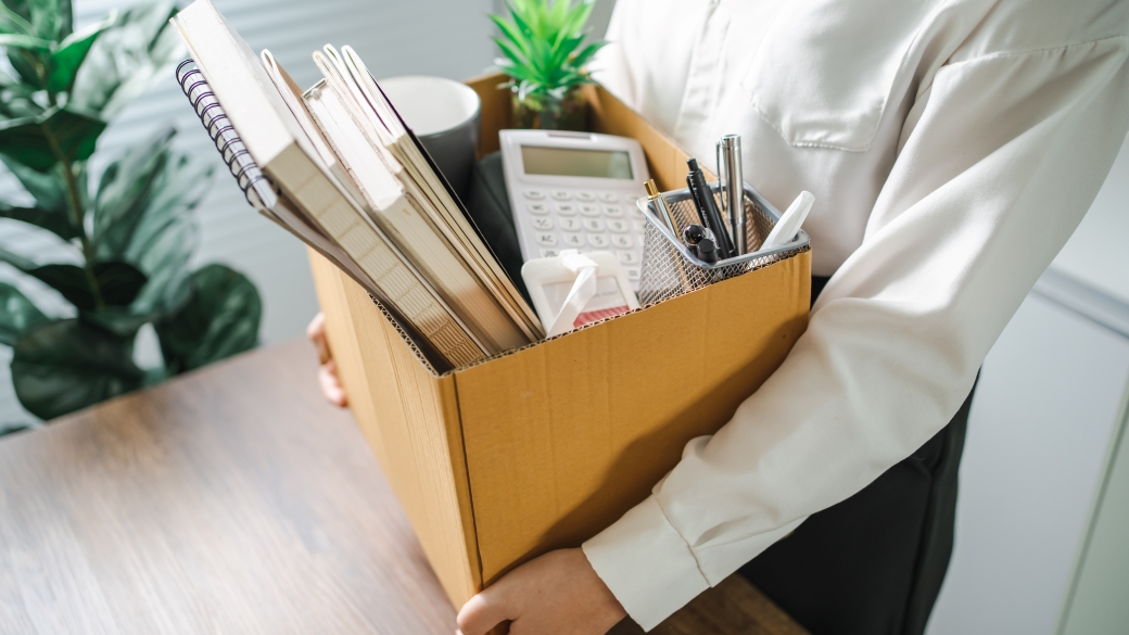 Foto: Eine Frau trägt im Büro eine Kiste mit ihren persönlichen Gegenständen vor sich her. Sie wurde entlassen.