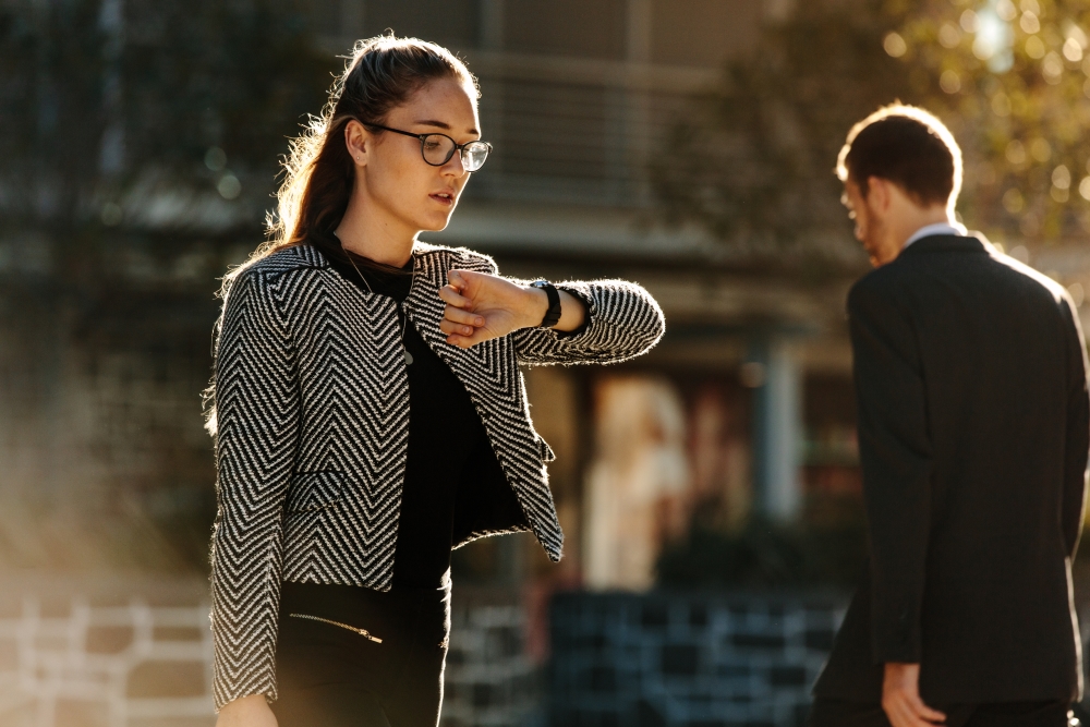 Foto: Eine Frau in Business-Kleidung steht im Sonnenschein und schaut nervös auf die Uhr.