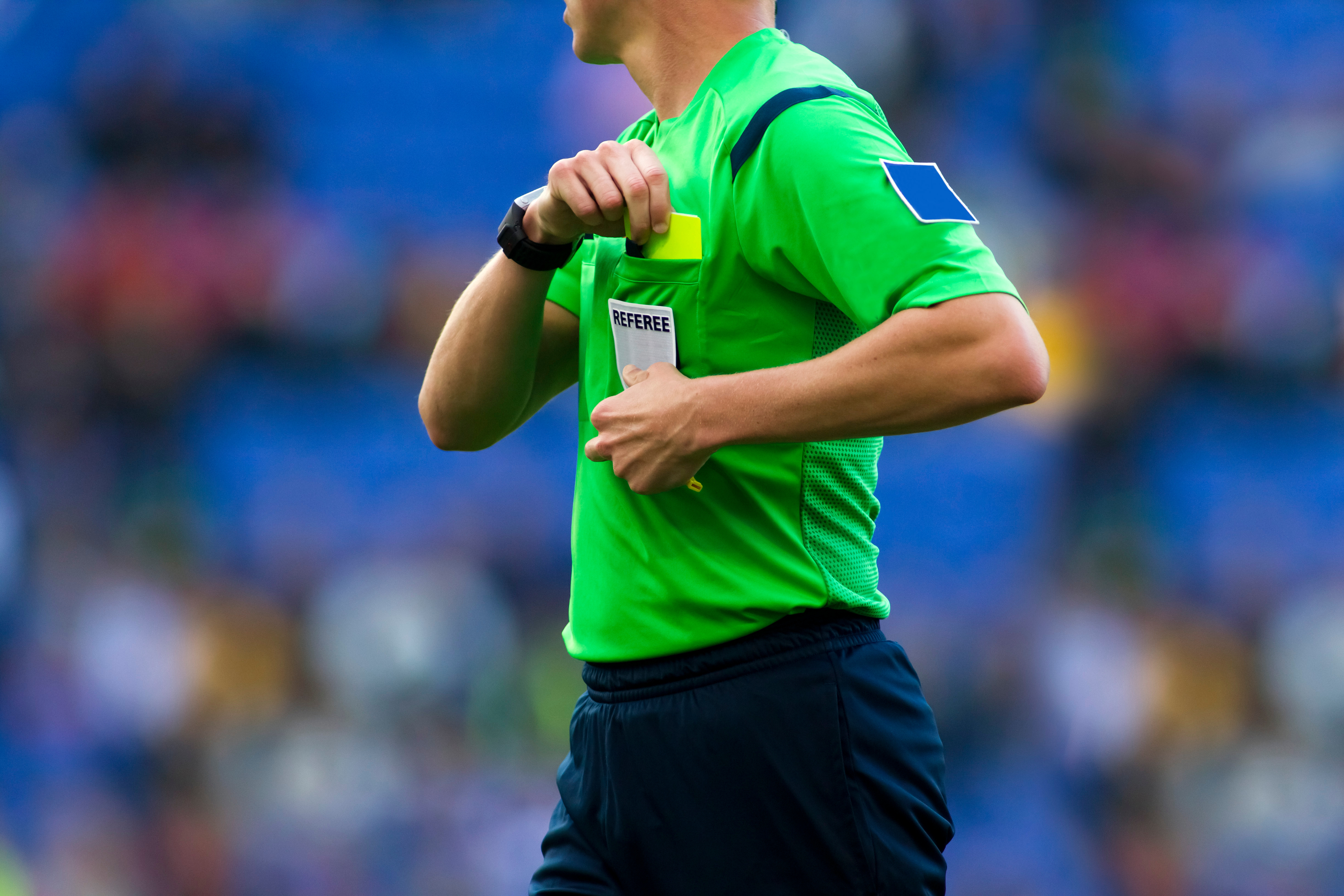 Foto: Ein Fußball-Schiedsrichter im hellgrünen Referee-Trikot bewegt seine rechte Hand zur linken Brusttasche. Darin befindet sich die Gelbe Karte.