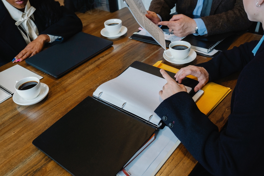 Foto: Drei Personen im Anzug sitzen mit einer Tasse Kaffe und Aktenordnern an einem Konferenztisch.