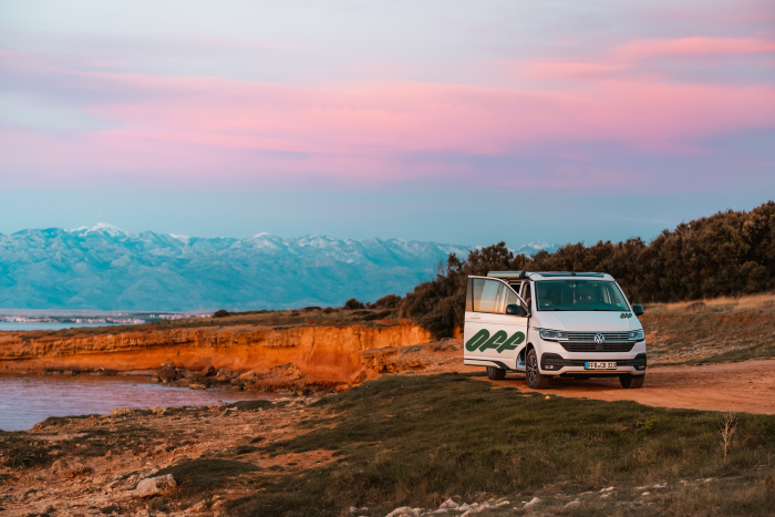 Foto: Ein Wohnmobil aus der Flotte von Off steht an einer Küste. Der Himmel ist Violett-Rosa gefärbt, es ist Sonnenaufgang.