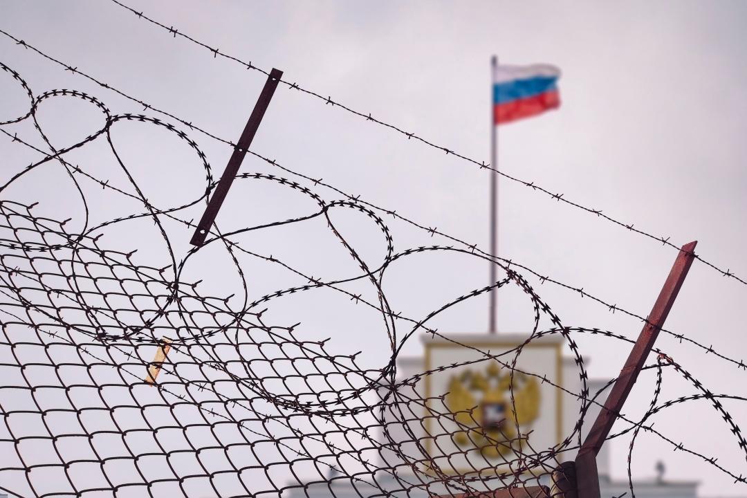 Foto: Stacheldrahtzaun. Im Hintergrund weht die russische Flagge auf einem Regierungsgebäude.