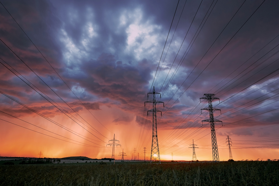 Foto: Eine Hochspannungsleistung mit Strommasten steht auf einem Feld im Sonnenuntergang. Darüber ziehen düstere Unwetterwolken auf.