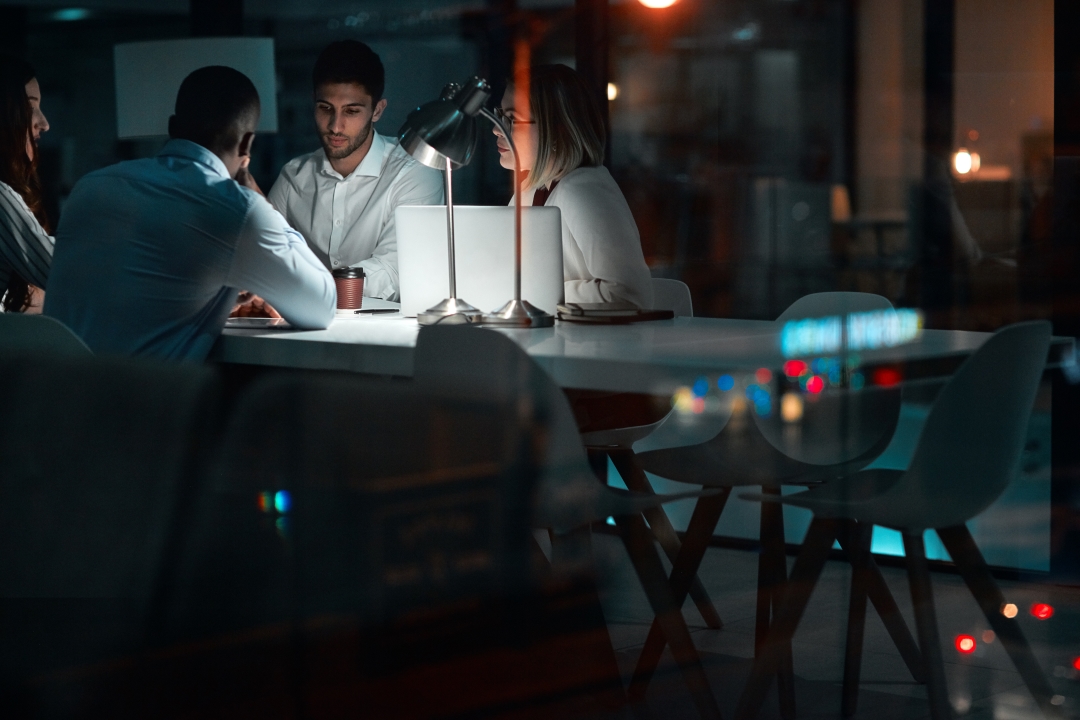 Photo: A group of start-up employees sit in a dark office at a conference table lit by a single desk lamp.