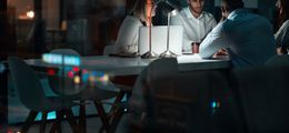 Photo: A group of start-up employees sit in a dark office at a conference table lit by a single desk lamp.