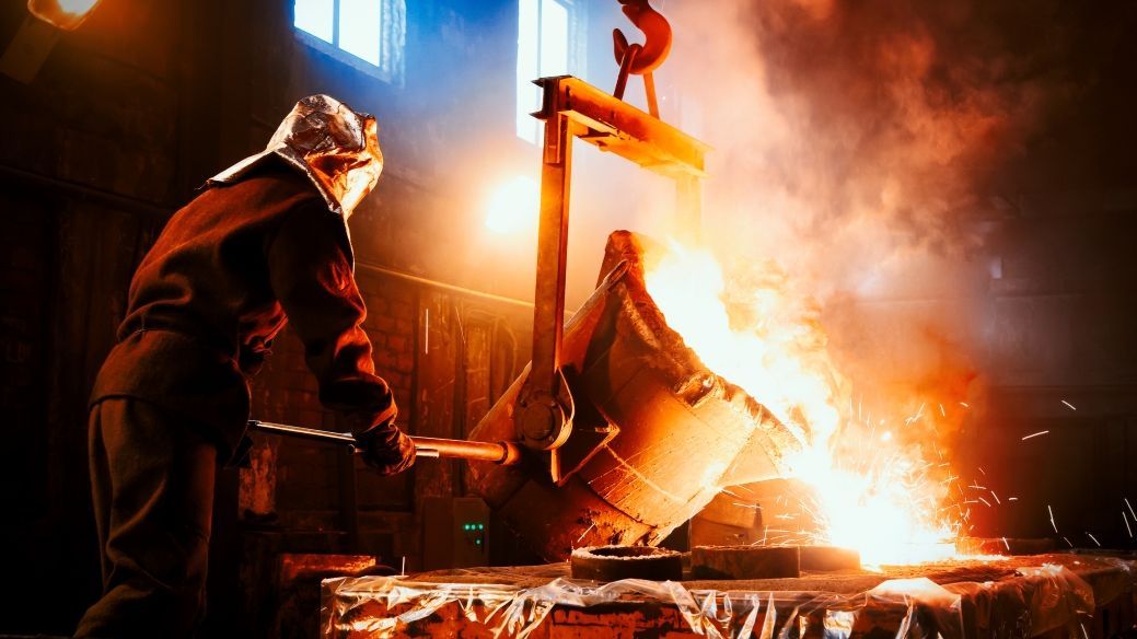 Foto: Ein Stahlarbeiter in feuerfester Kleidung steht in einem Stahlwerk. Er arbeitet an einem Bottich mit flüssigem Stahl, der gerade in einer Gießerei ausgekippt wird.