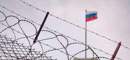 Foto: Stacheldrahtzaun. Im Hintergrund weht die russische Flagge auf einem Regierungsgebäude.